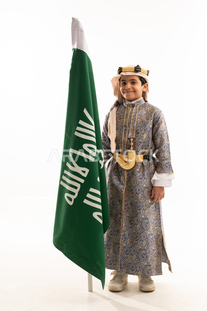 يوم بدينا 1727 م، النظر إلى الكاميرا بإيماءات الفخر، ارتداء أزياء المنطقة الوسطى في المناسبات الوطنية، بورتريه لولد عربي خليجي سعودي مبتسم يرتدي الدقلة والخنجر والعقال التقليدي الشعبي يقف بجانب علم تأسيس الدولة السعودية 22 فبراير، صورة لكامل الجسم، خلفية بيضاء