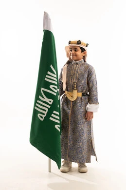 ارتداء أزياء المنطقة الوسطى في اليوم الوطني السعودي23سبتمبر، يوم بدينا 1727 م، الوقوف باستقامة بإيماءات الفخر، بورتريه لولد عربي خليجي سعودي مبتسم يرتدي الدقلة والخنجر والعقال التقليدي الشعبي يقف بجانب علم تأسيس الدولة السعودية 22 فبراير، صورة لكامل الجسم، خلفية بيضاء