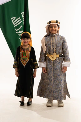 Showing off Saudi heritage, the anniversary of the founding of the first Saudi state, February 22, the day of our beginnings, 1727 AD, wearing traditional folk costumes of the southern and central regions, portrait of a smiling Saudi Gulf Arab boy and girl holding the founding flag, full body, white background