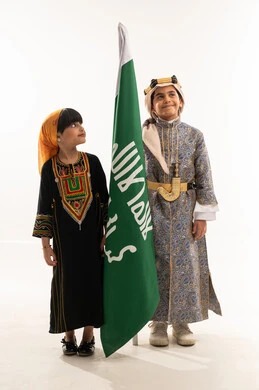 Wearing traditional folk costumes of the southern and central regions, showing off Saudi heritage, commemorating the founding of the first Saudi state, February 22, the day of our beginnings, 1727 AD, portrait of a smiling Saudi Arabian Gulf boy and girl holding the founding flag, full body, white background