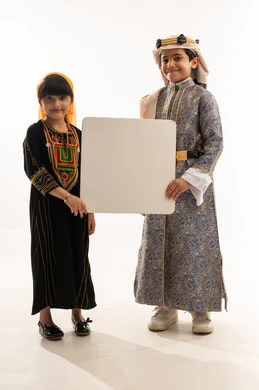 Reviving Saudi heritage, commemorating the founding of the first Saudi state, February 22, looking at the camera with gestures of pleasure, the day we began 1727 AD, wearing traditional folk costumes, portrait of a smiling Saudi Arabian Gulf boy and girl holding a large empty white mockup, full body, white background