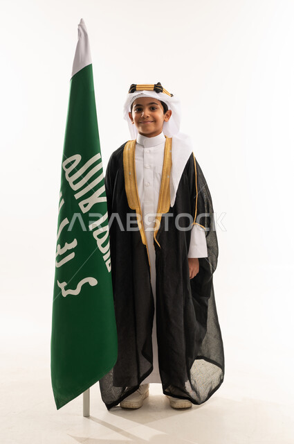النظر إلى الكاميرا بفخر بالانتماء للوطن، ارتداء أزياء المنطقة الشرقية في اليوم الوطني السعودي23سبتمبر، يوم بدينا 1727 م، بورتريه لولد عربي خليجي سعودي مبتسم يرتدي البشت والغترة وعقال التأسيس يمسك علم تأسيس الدولة السعودية 22 فبراير، صورة لكامل الجسم، خلفية بيضاء