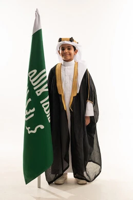Looking at the camera with pride of belonging to the homeland, wearing the Eastern Province costumes on national occasions, the day of our beginnings 1727 AD, a portrait of a smiling Saudi Gulf Arab boy wearing a bisht, ghutra and founding agal holding the founding flag of the Saudi state, February 22, a full body photo, white background