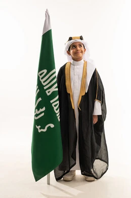 الوقوف باستقامة والنظر إلى الاعلى بفخر، ارتداء أزياء المنطقة الشرقية في المناسبات الوطنية، يوم بدينا 1727 م، بورتريه لولد عربي خليجي سعودي مبتسم يرتدي البشت والغترة وعقال التأسيس يمسك علم تأسيس الدولة السعودية 22 فبراير، صورة لكامل الجسم، خلفية بيضاء