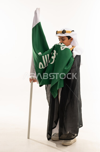 تربية الأبناء على حب الوطن، ارتداء أزياء المنطقة الشرقية في اليوم الوطني 23سبتمبر، يوم بدينا 1727 م، بورتريه لولد عربي خليجي سعودي مبتسم يرتدي البشت والغترة وعقال التأسيس يقوم بتقبيل علم تأسيس الدولة السعودية 22 فبراير، صورة لكامل الجسم، خلفية بيضاء