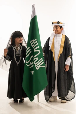 تربية الأبناء على الحب والولاء للوطن، زي تراثي سعودي للمناسبات الوطنية، يوم بدينا 1727 م، بورتريه لولد وبنت عربيين خليجيين سعوديين مبتسمين يمسكان علم تأسيس الدولة السعودية الأولى 22 فبراير، ، الجسم بالطول الكامل، خلفية بيضاء