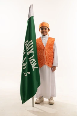 النظر إلى الكاميرا بإيماءات التباهي والفخر، ارتداء أزياء المنطقة الغربية في الاحتفالات والمناسبات الوطنية، يوم بدينا 1727 م، بورتريه لولد عربي خليجي سعودي مبتسم يرتدي السديري الشعبي يمسك علم تأسيس الدولة السعودية الأولى 22 فبراير، صورة الجسم بالكامل, خلفية بيضاء