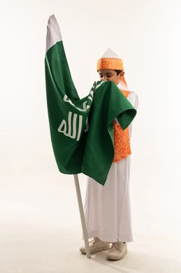 Raising children to love their country, wearing the Western Region's costumes on a national occasion, the Day of Our Beginning 1727 AD, portrait of a smiling Saudi Arabian Gulf boy wearing the traditional Sudairi, kissing the flag of the founding of the first Saudi state, February 22, full body image, white background