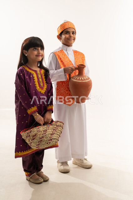 Popular handicrafts, looking at the camera with happy gestures, Saudi traditional costume on National Day September 23, the founding of the first Saudi state February 22, the day we began 1727 AD, a portrait of a smiling Saudi Arabian Gulf boy and girl holding a pottery pot and a straw basket, full body, white background