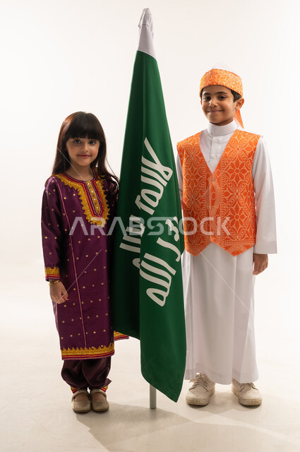 زي تراثي في اليوم الوطني السعودي 23سبتمبر، إظهار الحب والولاء للوطن، ذكرى تأسيس الدولة السعودية الأولى 22 فبراير، يوم بدينا 1727 م، بورتريه لولد وبنت عربيين خليجيين سعوديين مبتسمين يمسكان علم التأسيس، الجسم بالطول الكامل، خلفية بيضاء