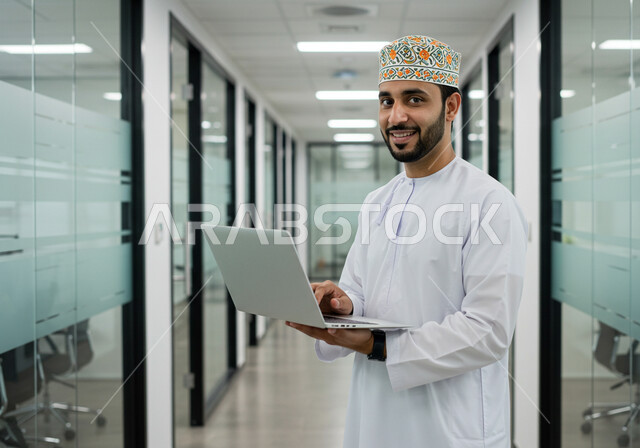 ادارة وانجاز المهام باستخدام جهاز تقني حديث ومتطور, رجل عربي خليجي عماني مبتسم يرتدي الكمة والدشداشة التقليدية يعمل على الحاسوب المحمول في مقر الشركة, ايماءات وتعابير السرور والسعادة، مهن ووظائف مكتبية عمانية 