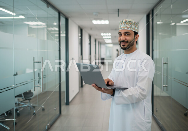 ادارة وانجاز المهام باستخدام جهاز تقني حديث ومتطور, رجل عربي خليجي عماني مبتسم يرتدي الكمة والدشداشة التقليدية يعمل على الحاسوب المحمول في مقر الشركة, ايماءات وتعابير السرور والسعادة، مهن ووظائف مكتبية عمانية 
