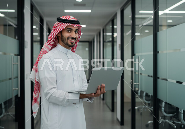 Managing and completing tasks using a modern and advanced technical device, a smiling Saudi Arabian Gulf man wearing a traditional shemagh and thobe working on a laptop at the company headquarters, gestures and expressions of pleasure and happiness, Saudi office jobs and professions