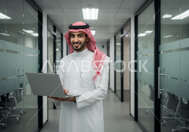 ادارة وانجاز المهام باستخدام جهاز تقني حديث ومتطور, رجل عربي خليجي سعودي مبتسم يرتدي الشماغ والثوب التقليدي يعمل على الحاسوب المحمول في مقر الشركة, ايماءات وتعابير السرور والسعادة، مهن ووظائف مكتبية سعودية