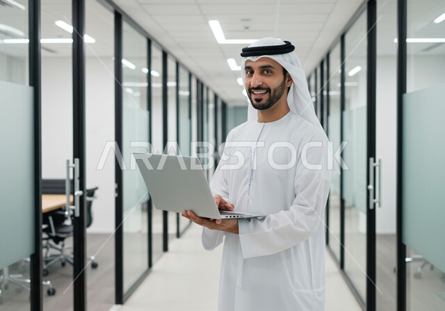 ادارة وانجاز المهام باستخدام جهاز تقني حديث ومتطور, رجل عربي خليجي اماراتي مبتسم يرتدي الغترة والكندورة التقليدية يعمل على الحاسوب المحمول في مقر الشركة, ايماءات وتعابير السرور والسعادة، مهن ووظائف مكتبية اماراتية