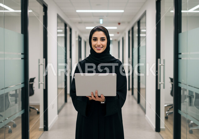  مهن ووظائف نسائية مكتبية سعودية، ادارة وانجاز المهام باستخدام جهاز تقني حديث ومتطور, إمرأة عربية خليجية سعودية مبتسمة ترتدي الحجاب والعباءة تعمل على الحاسوب المحمول في مقر الشركة, ايماءات وتعابير السرور والسعادة
