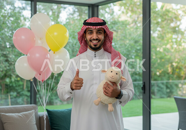 ايماءات البهجة والفرح والسعادة, رجل عربي خليجي سعودي مبتسم يرتدي الثوب التقليدي والشماغ و يحمل بيده دمية على شكل خاروف عيد الاضحى المبارك, أعياد ومناسبات اسلامية, الاحتفال بالعيد السعيد في المنزل