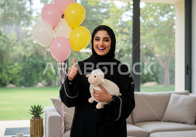 ايماءات البهجة والفرح والسعادة, إمرأة عربية خليجية سعودية مبتسمة ترتدي الحجاب والعباءة تحمل بيدها دمية على شكل خاروف عيد الاضحى المبارك, أعياد ومناسبات اسلامية, الاحتفال بالعيد السعيد في المنزل