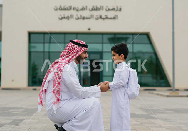 التحفيز المعنوي للأبناء في أول أيام الدوام المدرسي، إلقاء عبارات التشجيع الداعمة وتقديم الدعم النفسي، موسم العودة إلى المدارس بالمملكة العربية السعودية، مفهوم الأبوة والحنان، اب عربي خليجي سعودي يرتدي الشماغ والثوب التقليدي ويقف برفقة ابنه