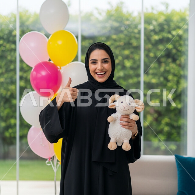 Gestures of joy, happiness and happiness, a smiling Saudi Arabian Gulf woman wearing a hijab and abaya holding a doll in the shape of a sheep for Eid al-Adha, Islamic holidays and occasions, celebrating the happy Eid at home