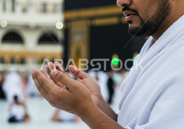 Performing religious duties and worship and getting closer to God, a close-up image of the hands of a Muslim man wearing the ihram clothes and raising his hands in supplication and prayer in front of the Holy Kaaba, Muslims heading to perform the Hajj and Umrah rituals