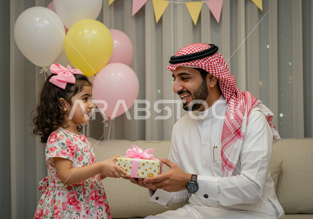  اجواء العائلة السعودية في الاعياد والمناسبات السعيدة، بهجه وفرحة تجمع الاسرة، ايماءات وتعابير السرور والسعادة، مفهوم صلة الرحم، رجل عربي خليجي سعودي يرتدي الشماغ والثوب التقليدي يقوم بتقديم الهدية لابنته