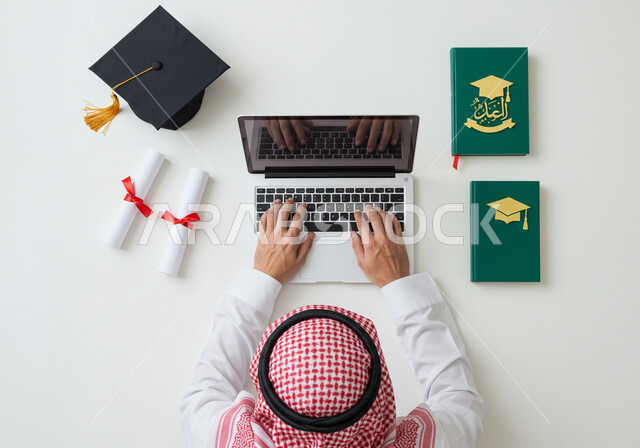 Education in Saudi Arabia, concept of diligence and excellence, vertical overhead portrait of a Saudi Arabian Gulf man wearing traditional thobe and shemagh writing a research paper on a laptop with a graduation cap and certificate next to him, white background