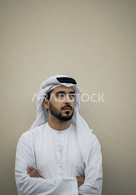 النظر الى مكان ما بايماءات الثقة بالنفس، الاهتمام بالمظهر الخارجي, مفهوم الأناقة والرجولة, بورتريه لرجل عربي خليجي اماراتي يرتدي الكندورة يقف بأيدي متقاطعة، التقاط الصور الشخصية للوثائق الرسمية، خلفية بيضاء