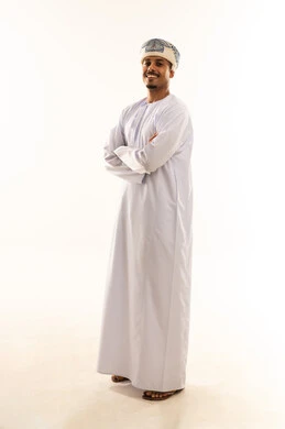 مفهوم الرجولة والوقار، الاهتمام بالمظهر الخارجي، تعابير الثقة بالنفس، بورتريه لرجل عربي خليجي عماني  مبتسم يرتدي الدشداشة والعمامة يقف بايدي متقاطعة ينظر الى الكاميرا بايماءات الثقه و السرور، صورة الجسم بالطول الكامل، خلفية بيضاء