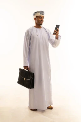 Hearing good news from relatives, browsing social media, chatting with family and friends, using modern technology, a portrait of a young Arab Gulf Omani man wearing a dishdasha and a turban holding a mobile phone and a briefcase, full-body, white background.