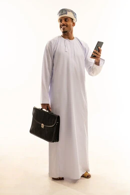 Browsing social media, chatting with family and friends, using a modern technological device, a portrait of a young Arab Gulf Omani man wearing a dishdasha and a turban holding a mobile phone and a briefcase looking at something, hearing good news from relatives, full-body, white background.