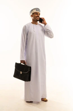 إيماءات السعادة بالتواصل مع الأصدقاء والأقارب، الاستمتاع بتبادل الأحاديث و الأخبار، إجراء مكالمة هاتفية صوتية عبر الجوال، بورترية من الجانب لرجل عربي  خليجي عماني  يرتدي الدشداشة و العمامة يمسك بيديه حقيبه العمل و يقوم بالتحدث عبر الهاتف المحمول، الجسم بالطول الكامل ، خلفية بيضاء