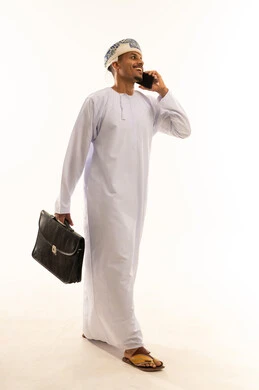 Enjoying exchanging conversations and news, making a voice call via mobile, a side portrait of an Arab Gulf Omani man wearing a dishdasha and a turban holding a briefcase in his hands and talking on a mobile phone, gestures of happiness in communicating with friends and relatives, full-body, white background.