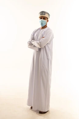 Attention to appearance, the concept of masculinity and dignity, expressions of self-confidence, a portrait of an Arab Gulf Omani man wearing a dishdasha, turban, and mask standing with arms crossed, full-body image, white background.