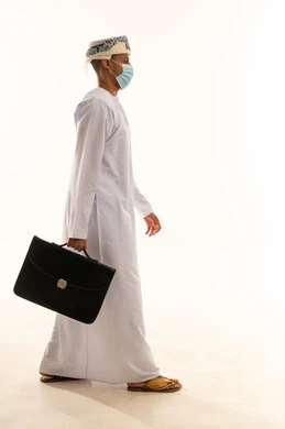 الذهاب إلى العمل بنشاط، بورتريه من الجانب لرجل عربي خليجي عماني يرتدي الدشداشة و العمامة و الكمامة يحمل بيده حقيبة يد يمشي إلى مكان ما ينظر أمامه بإيماءات الثقة بالنفس، الاهتمام بالمظهر الخارجي، مفهوم إداة الأعمال والمشاريع، الجسم بالطول الكامل، خلفيه بيضاء