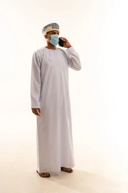 دردشات ومحادثات عبر الانترنت، إجراء المكالمات الصوتية، بورتريه لرجل عربي خليجي عماني يرتدي الدشداشة و العمامة و الكمامة يستخدم الهاتف المحمول، التواصل مع الأصدقاء عبر مواقع التواصل الاجتماعي، استخدام التكنولوجيا و التقنيات الحديثة, الطول الكامل للجسم، خلفية بيضاء