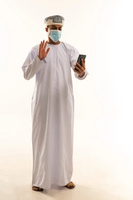 دردشة ومحادثات وتعارف، اجراء مكالمة فيديو، دمج التكنولوجيا في الحياة اليومية، استخدام جهاز تقني حديث ومتطور، بورتريه لرجل عربي خليجي عماني يرتدي الدشداشة و العمامة و الكمامة يقوم باستخدام الهاتف المحمول يرفع يده بايماءات القاء التحية والسلام، الجسم بالطول الكامل، خلفية بيضاء