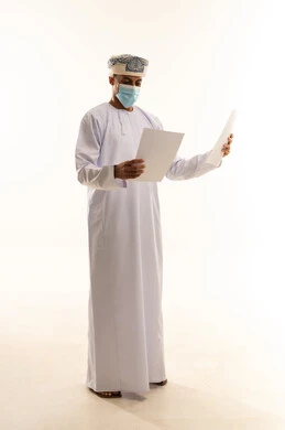 ايماءات الاندماج و التركيز، اعداد التقارير الخاصة بالعمل، بورتريه لشاب عربي خليجي عماني يرتدي الدشداشة و العمامة و الكمامة يحمل بيده مجموعة من الاوراق البيضاء، مراجعة ومتابعة الخطط والمشاريع المكتبية ، الجسم بالطول الكامل ، خلفية بيضاء