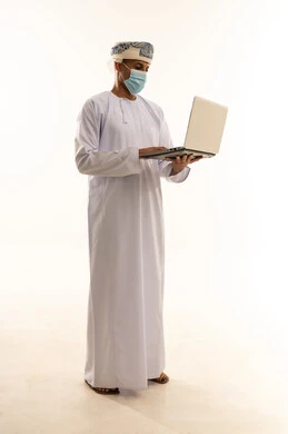دمج التقنية والتكنولوجيا الحديثة في الحياة اليومية، تصفح مواقع ووسائل التواصل الاجتماعي ، بورتريه لرجل عربي خليجي عماني يرتدي الدشداشة و العمامة و الكمامة  يقوم بإجراء مكالمة فيديو عبر جهاز الحاسوب المحمول, الجسم بالطول الكامل، خلفية بيضاء