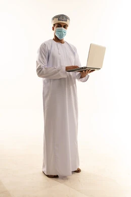 Using modern technologies to communicate with customers, a portrait of an Arab Gulf Omani man wearing a dishdasha, turban, and mask conducting a video call via a laptop, looking at the camera, full body, white background.