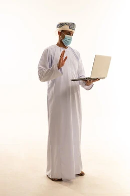 Making a video call via a laptop, using modern technologies to communicate with clients, a portrait of an Arab Gulf Omani man wearing a dishdasha, turban, and mask, raising his hands in gestures of greeting and peace, full-body, white background.