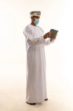 Watching enjoyable movies and series, browsing social media, chatting with family and friends, a portrait of an Arab Gulf Omani man wearing a dishdasha, turban, and mask holding a tablet in his hand, making a video call via the tablet, white background.