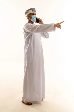 الاشارة الى شي ما ، الدردشة والتواصل مع الأهل والأصدقاء وسماع أخبارهم، بورتريه من الجانب لرجل عربي خليجي عماني يرتدي الدشداشة و العمامة و الكمامة يقوم بإجراء مكالمة هاتفية عبر الهاتف المحمول, استخدام جهاز تقني حديث, الجسم بالطول الكامل، خلفية بيضاء