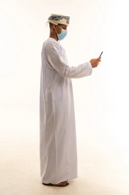 استخدام جهاز تقني حديث ومتطور, التسلية والترفيه في وقت الفراغ, تصفح وسائل التواصل الاجتماعي, بورتريه من الجانب لرجل عربي خليجي عماني يرتدي الدشداشة و العمامة و الكمامة يقوم باستعمال الهاتف المحمول, الجسم بالطول الكامل, خلفية بيضاء