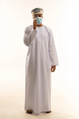 توضيح الصورة والرؤية عن قرب، مفهوم الفحص والاستكشاف، بورتريه لرجل عربي خليجي عماني يرتدي الدشداشة والعمامة و الكمامة يحمل بيده عدسة مكبرة يضعها على عينه بإيماءات التركيز والتمعن، خلفية بيضاء