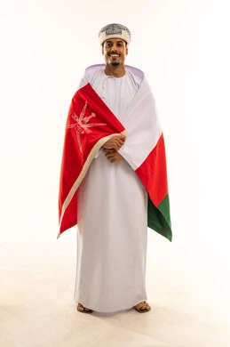 الاحتفال بذكرى اليوم الوطني العماني، النظر الى الكاميرا بإيماءات السرور و الفرح, الاعتزاز بالانتماء للهوية، الفخر بالرموز والراية العمانية، بورتريه لشاب عربي خليجي عماني يرتدي العمامة والدشداشة يضع العلم على اكتافه، مفهوم حب الوطن، الجسم بالطول الكامل، خلفية بيضاء