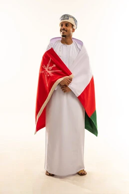 مفهوم حب الوطن، الاعتزاز بالانتماء للهوية، النظر شيء ما، الاحتفال بذكرى اليوم الوطني العماني، الفخر بالرموز والراية العمانية، بورتريه لشاب عربي خليجي عماني يرتدي العمامة والدشداشة  يضع العلم على اكتافه، الجسم بالطول الكامل، خلفية بيضاء