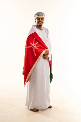 Looking at the camera with gestures of joy and happiness, pride in belonging to the identity, celebrating the anniversary of Oman's National Day, pride in the symbols and the Omani flag, a portrait of a young Arab Gulf Omani man wearing a turban and a dishdasha, draping the flag over his shoulders, the concept of love for the homeland, full-body shot, white background.