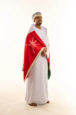 Pride in belonging to the identity, looking at something, celebrating the anniversary of Oman's National Day, pride in the symbols and the Omani flag, a portrait of a young Arab Gulf Omani man wearing a turban and a dishdasha, draping the flag over his shoulders, the concept of love for the homeland, full-body, white background.