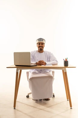 العمل في المكاتب والشركات العمانية، دمج التقنيات الحديثة بالمجال الإداري والمكتبية، بورتريه لرجل عربي خليجي عماني يرتدي الدشداشه والعمامه يجلس خلف طاولة المكتب امامه جهاز الحاسوب المحمول و يقوم بالنظر الكاميرا بايماءات السعادة و السرور، خلفية بيضاء